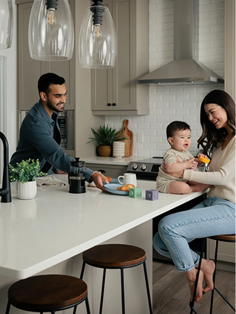 A man and woman sit at a kitchen island, smiling at a baby resting in the woman's arms.